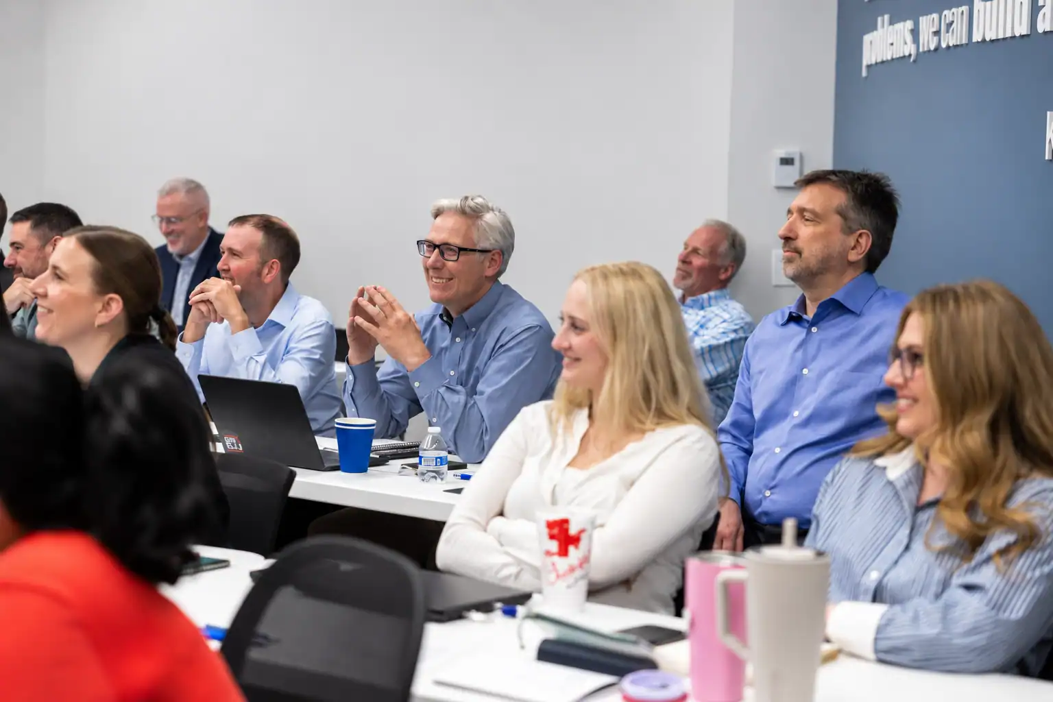 Group of diverse professionals attending a conference, sitting at tables, listening attentively, some smiling, in a modern conference room.
