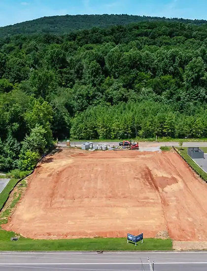 A cleared and leveled plot of land with red soil, ready for construction, surrounded by lush green trees and a forested hillside in the background.