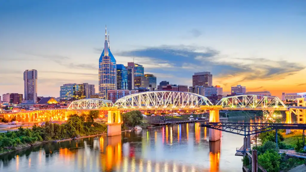Nashville skyline with the AT&T Building and a bridge over the Cumberland River during sunset, city lights beginning to glow.