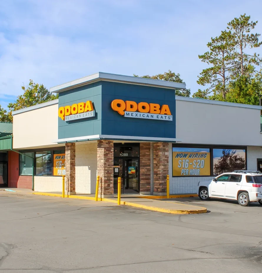 Exterior view of QDOBA Mexican Eats restaurant with parking lot and cars, clear sky, and surrounding trees.