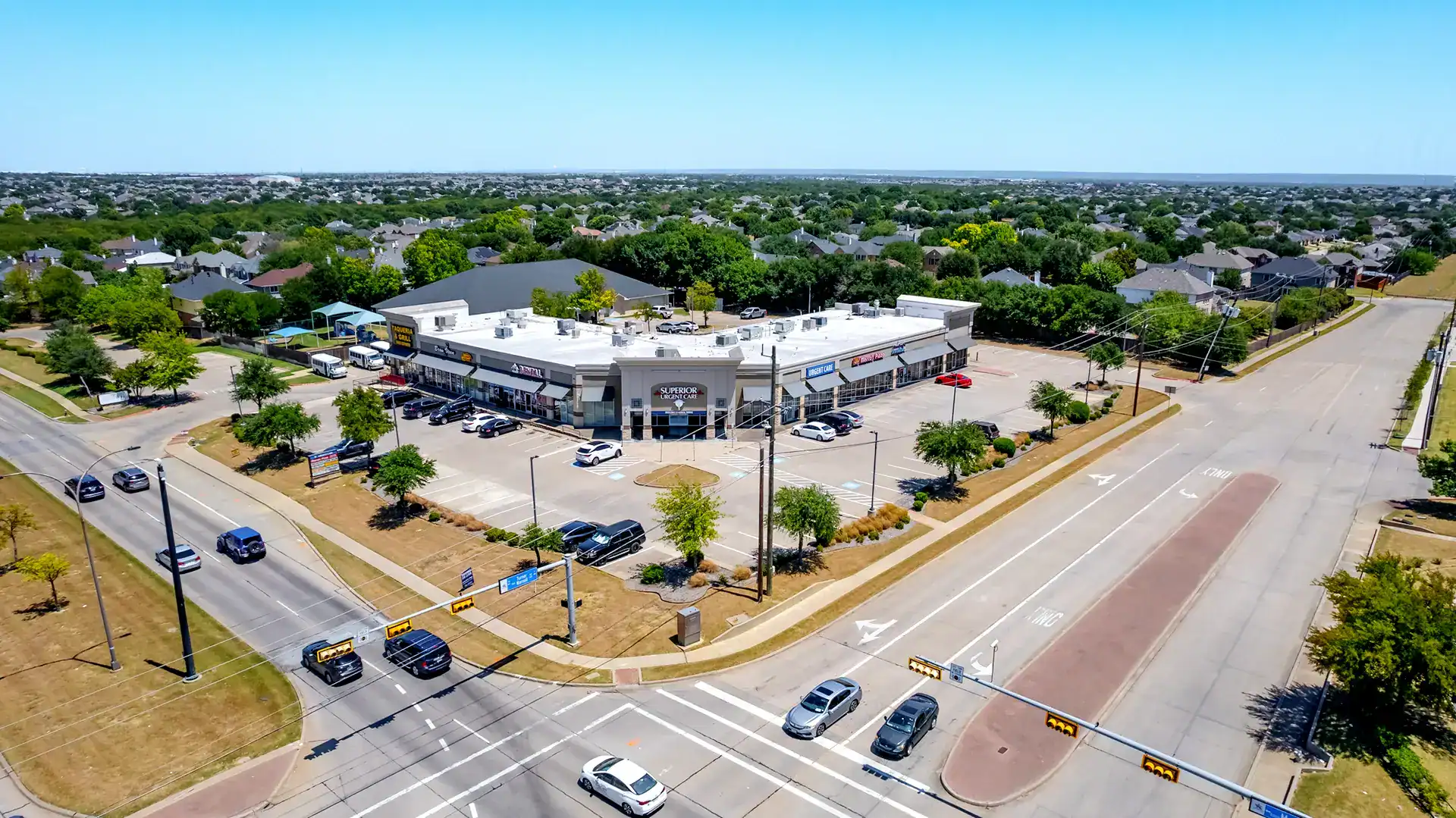 Aerial view of a shopping center with parking lot, surrounded by trees and roads in a suburban area.