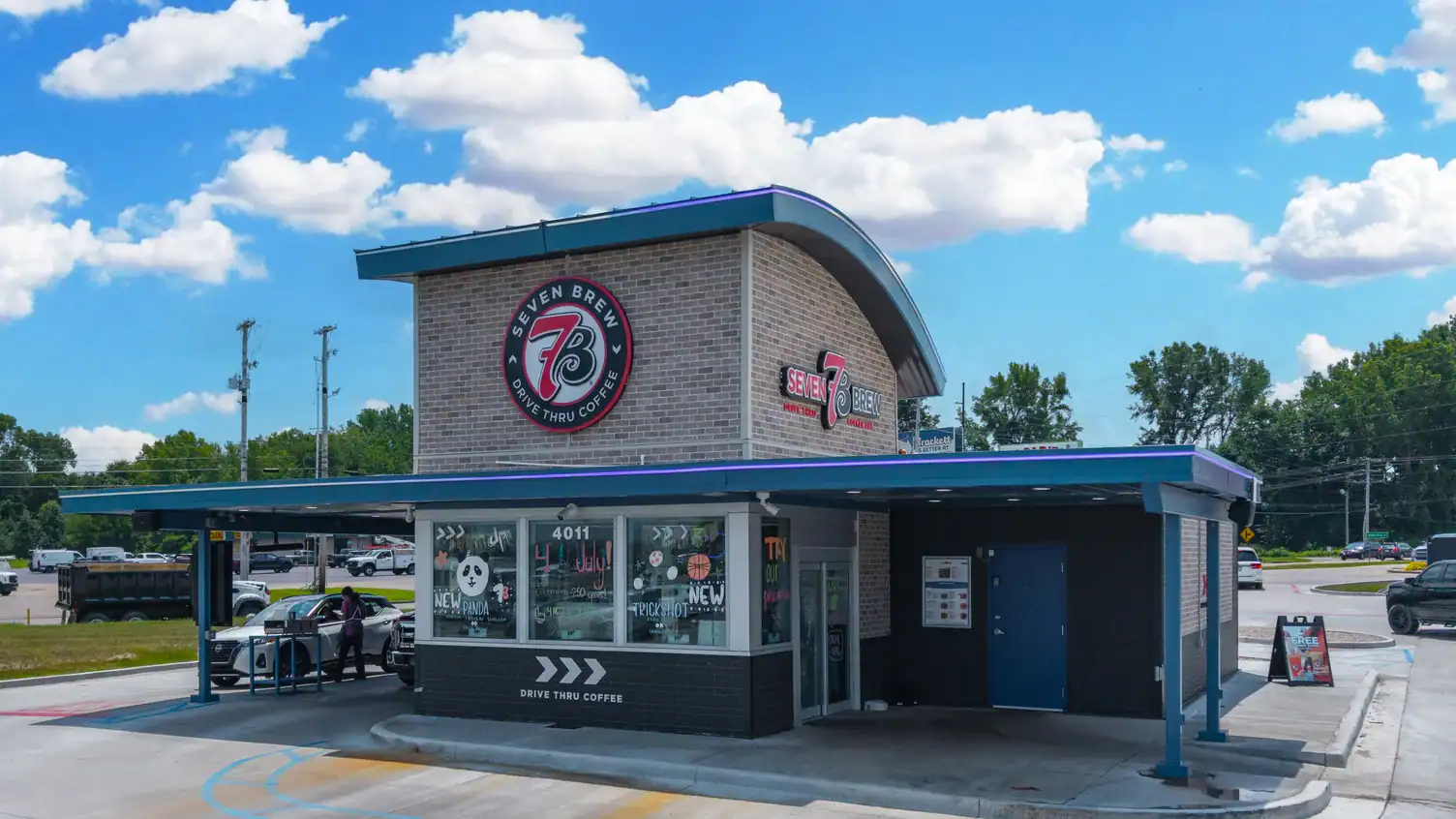 A modern coffee shop building with a curved roof and large windows, located in a parking lot on a sunny day.