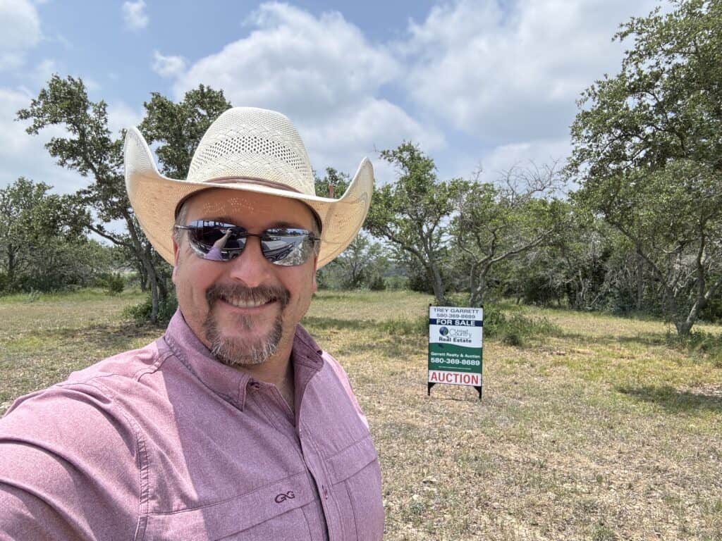 Man in cowboy hat and sunglasses standing near Garrett Realty & Auction sign in open field.