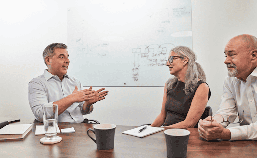 Three professionals seated around a conference table discussing ideas, with one man gesturing as a woman and another man listen in a bright meeting room.