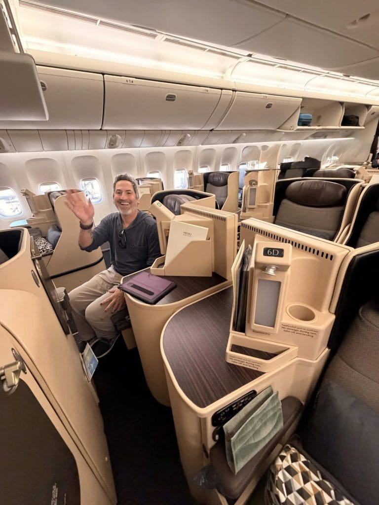 A smiling man waves from his comfortable business class seat on a modern airplane.