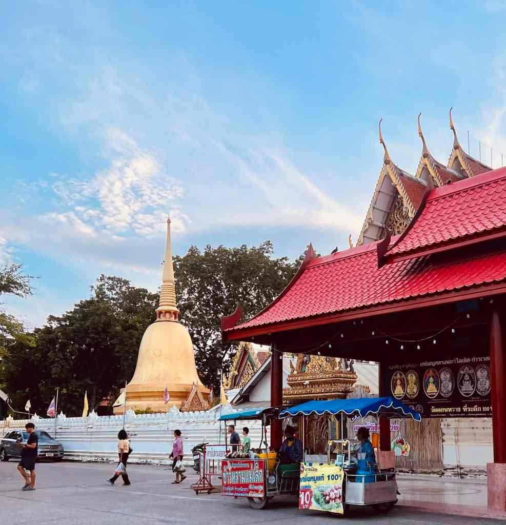 Temple scene featuring traditional Thai architecture with visitors and street vendors. Bright sky an.