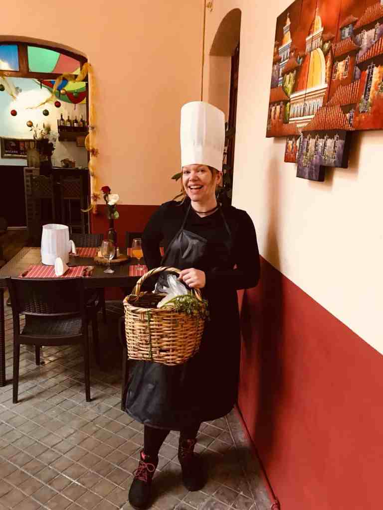 Chef wearing a tall white hat and black apron holding a basket, smiling in a warm, inviting restaura.