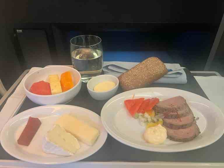 Airplane meal featuring beef, cheese, bread, and fresh fruit on tray table.