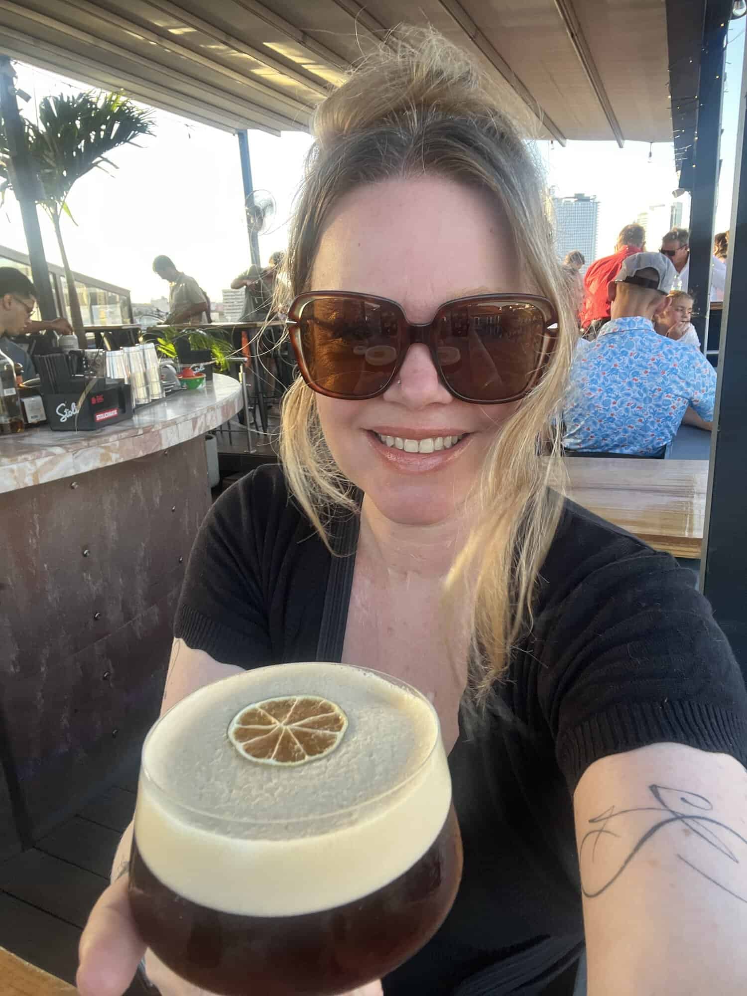 Woman with sunglasses holding a cocktail at a rooftop bar during travel.