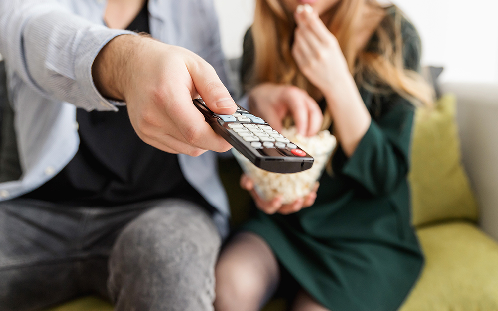 A couple enjoying a cozy night in, watching a movie and sharing popcorn with the man holding the remote control.