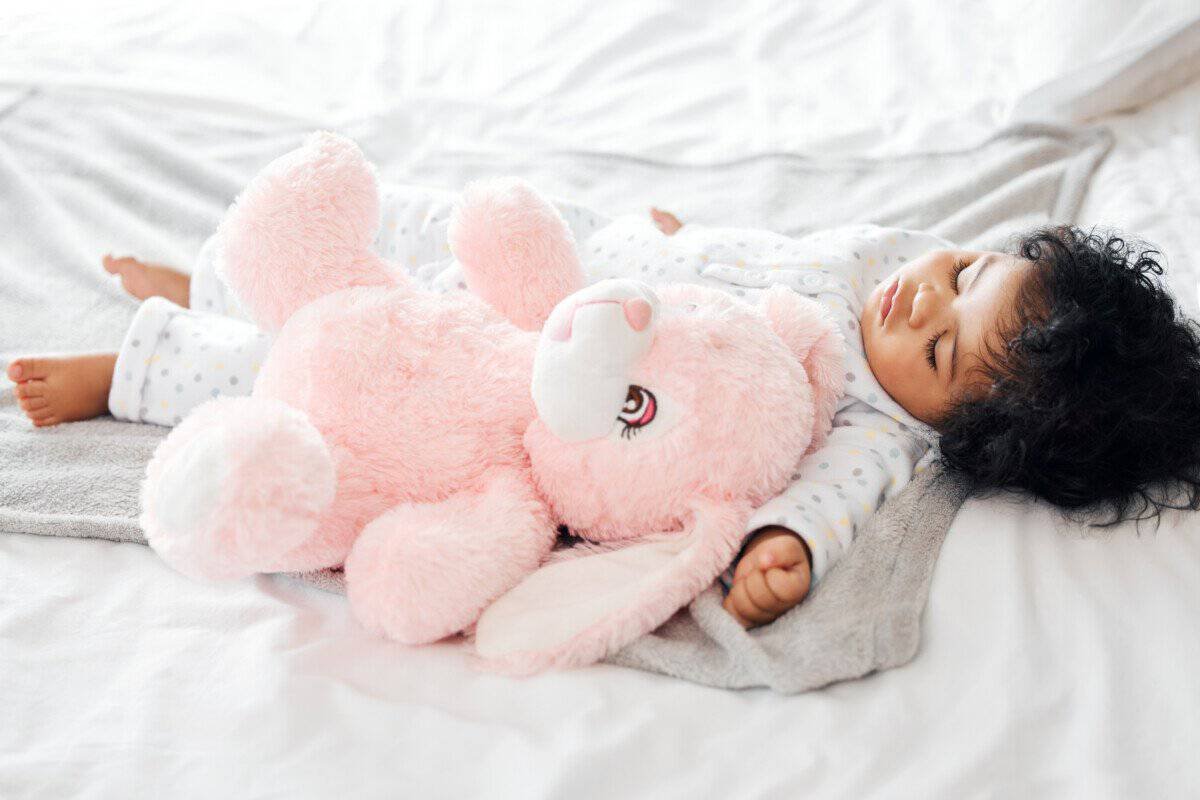 A toddler, curls riotous and dreams epic, slumbers in printed pyjamas while a sprawling pink bunny stands sentry atop the blankets.
