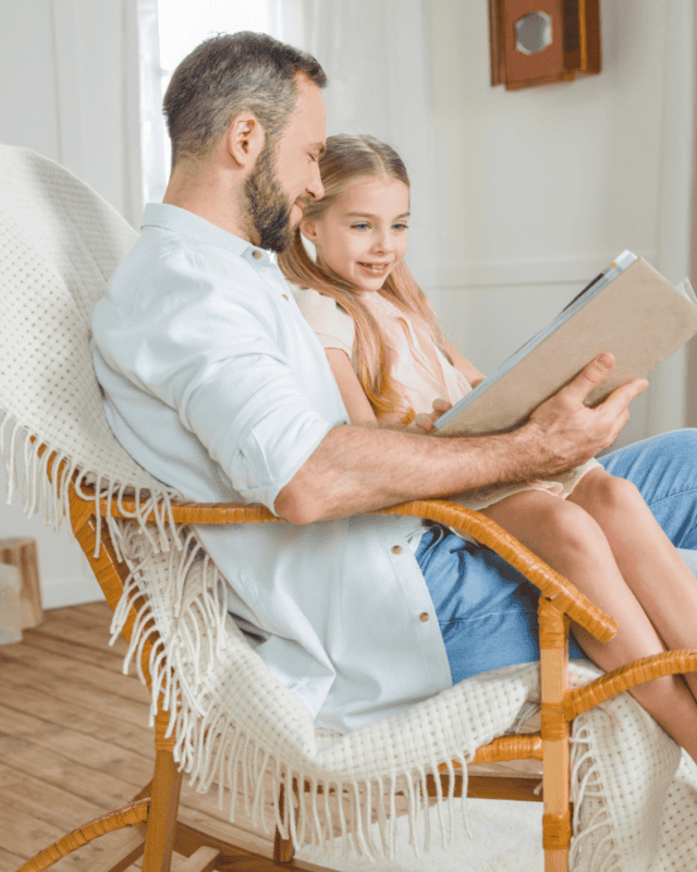 In a sunlit room, a man in wispy garb reads to a grinning girl on his lap; chair and blanket embroiled in utmost cosiness.