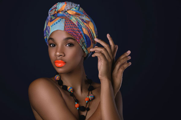 Vendeuse africaine avec un foulard coloré et un collier artisanal, pose élégante sur fond noir.