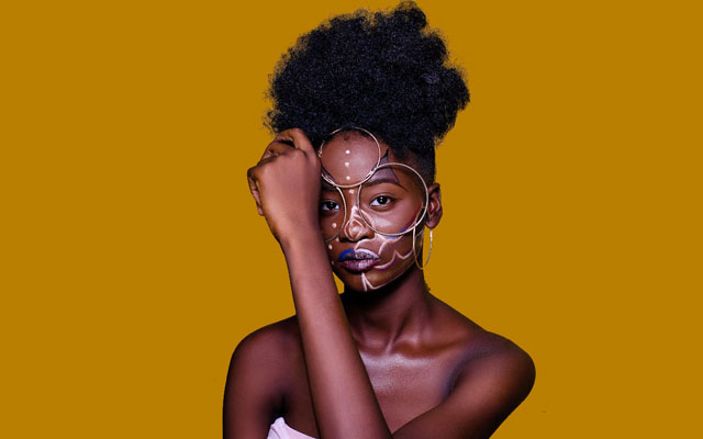 L'image montre une femme avec un maquillage artistique et des accessoires géométriques sur le visage, sur un fond jaune vif.