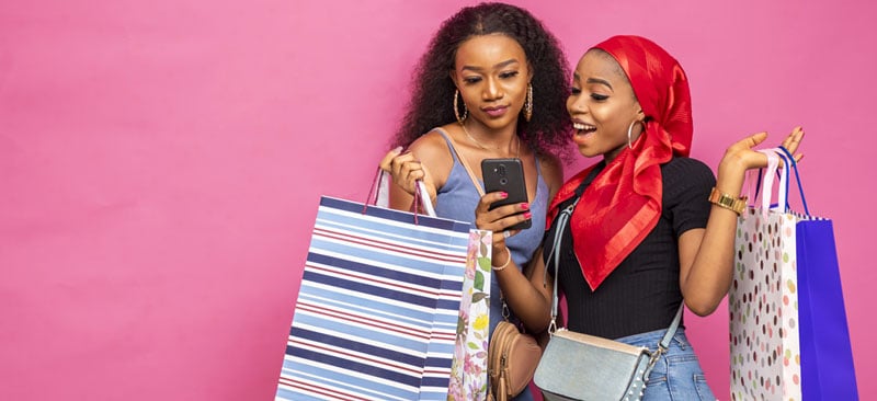 Deux femmes de shopping regardant leur téléphone avec des sacs colorés contre un fond rose.
