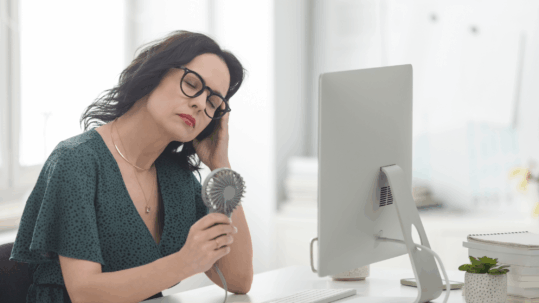 Woman sitting at a desk with a hand-held fan, looking tired or overheated, illustrating the emotional and physical symptoms of menopause like hot flashes, anxiety, and brain fog. Image relates to hormone therapy for women's emotional health.