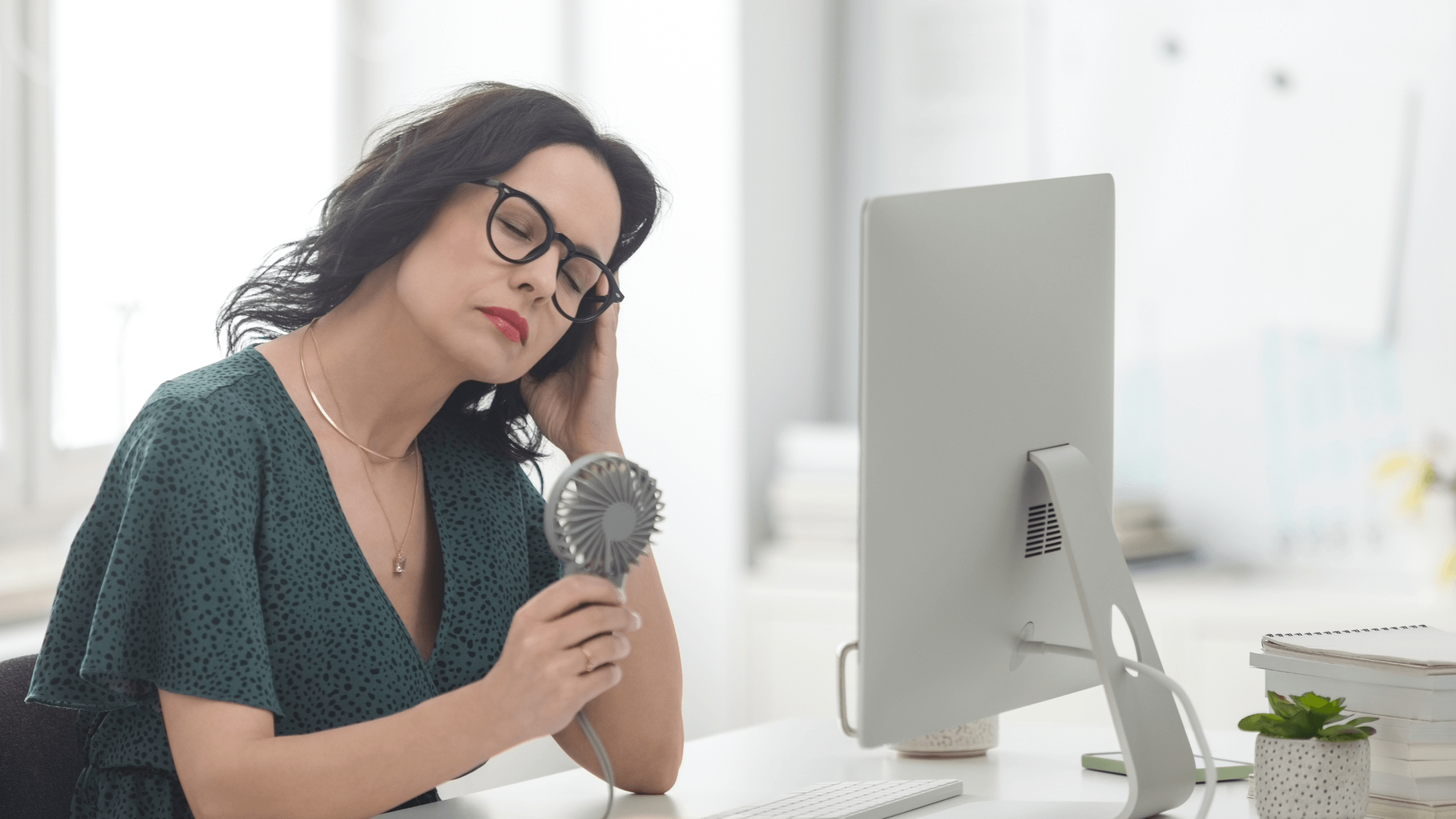 Woman sitting at a desk with a hand-held fan, looking tired or overheated, illustrating the emotional and physical symptoms of menopause like hot flashes, anxiety, and brain fog. Image relates to hormone therapy for women's emotional health.