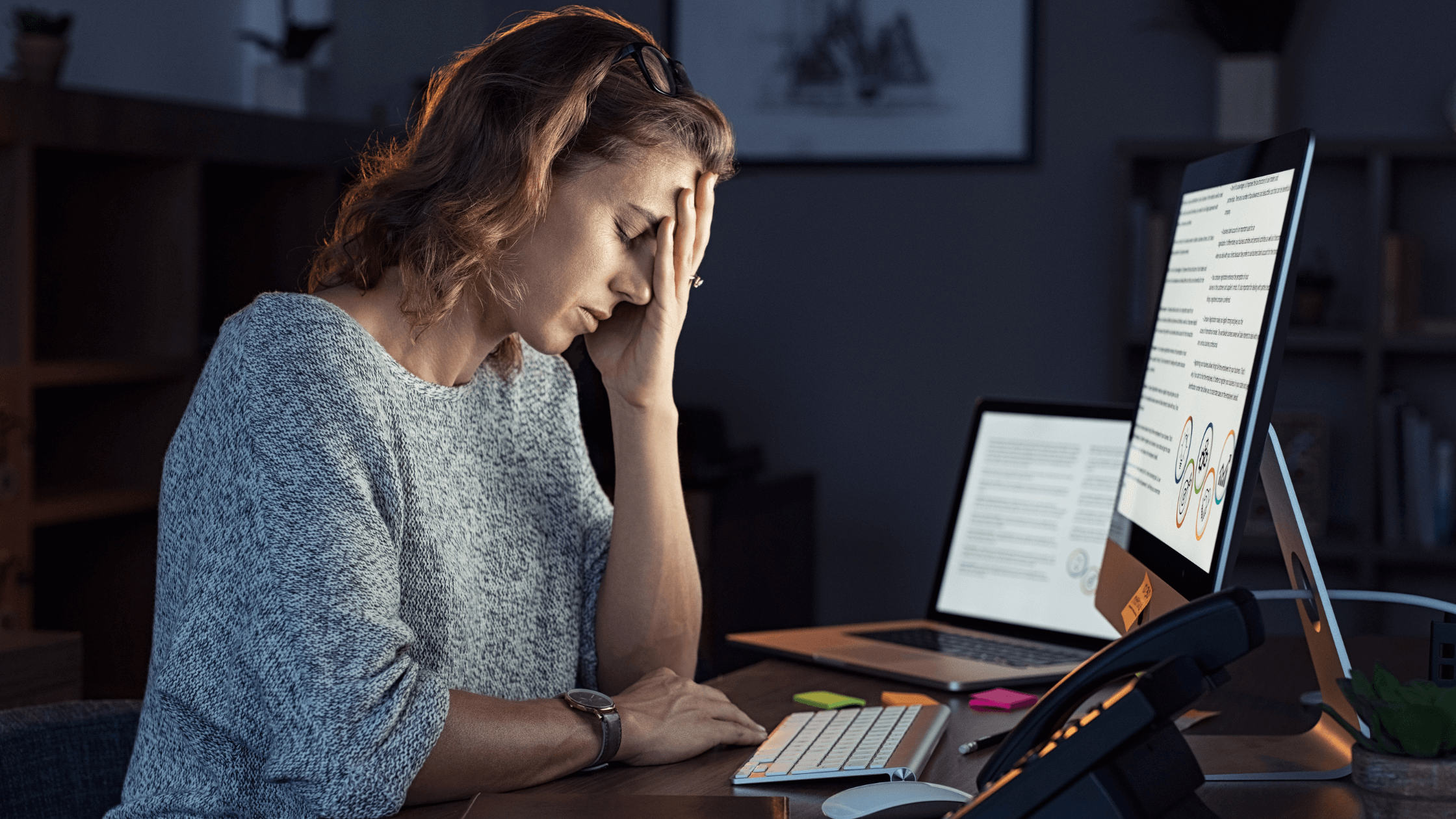 A distressed woman holding her head at a dimly lit desk, illustrating chronic fatigue, brain fog, and stress from hormonal instability during perimenopause, linking to hormone therapy and wellness in Colleyville, Fort Worth, or Southlake.