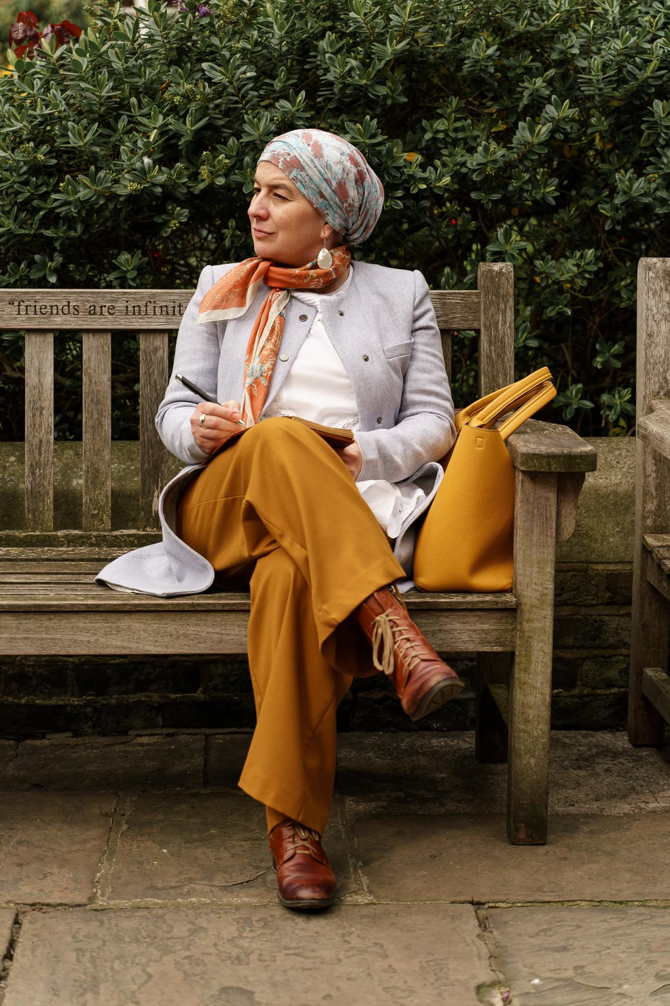 A confident woman in a stylish headscarf and mustard trousers sits on a wooden bench in London, writing, captured by a London brand photographer.