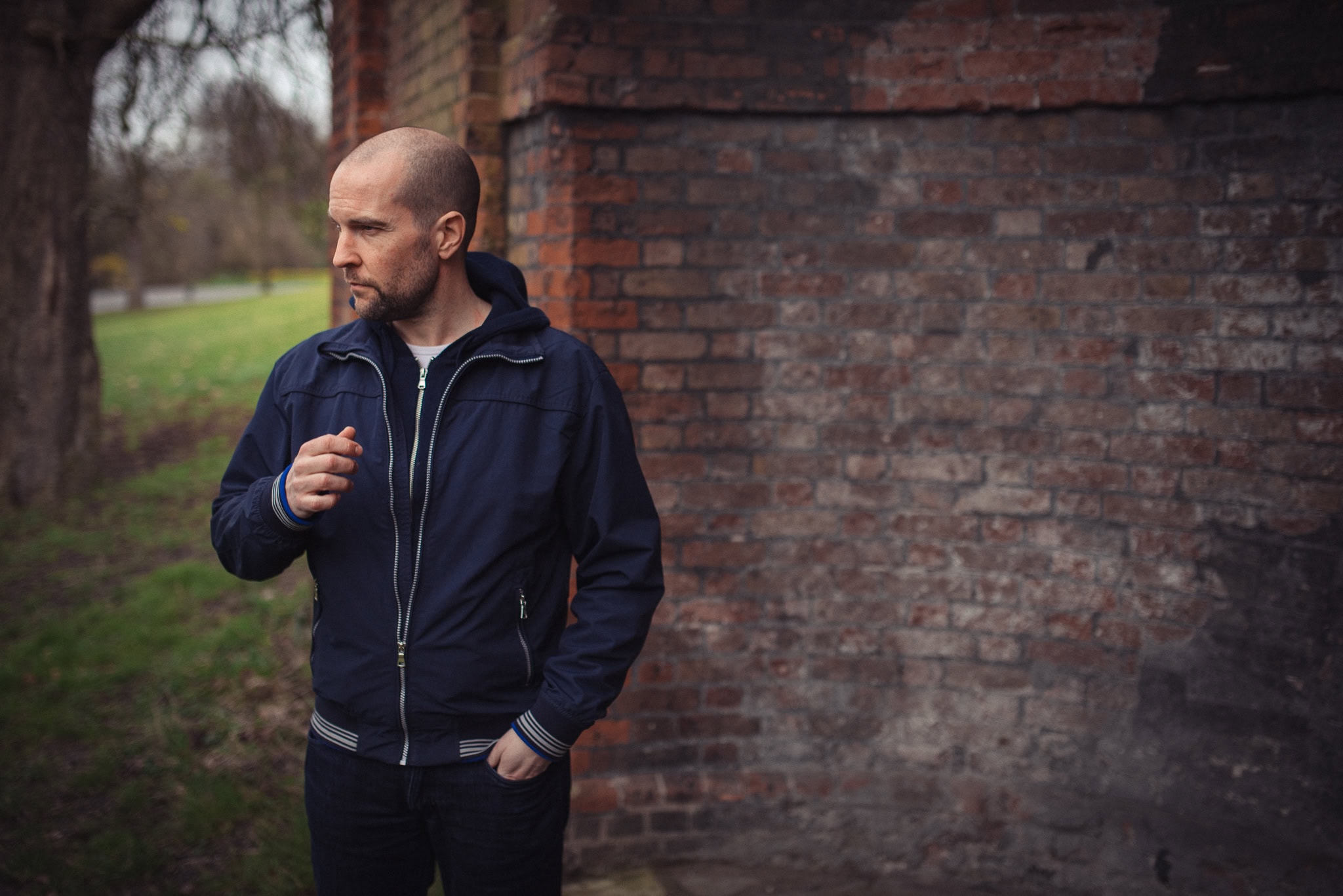 Bald man in navy jacket standing by a brick wall in a park