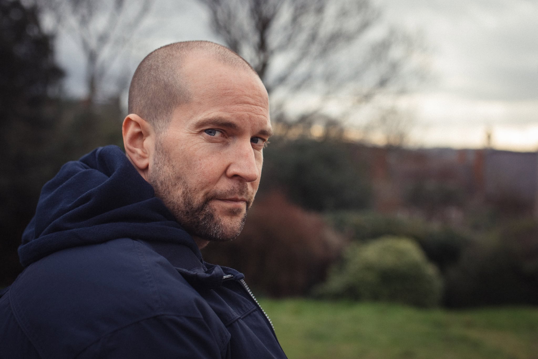 Clive Maxheath, a man with a shaved head and beard, looking over his shoulder in a natural outdoor setting, captured by a London brand photographer.