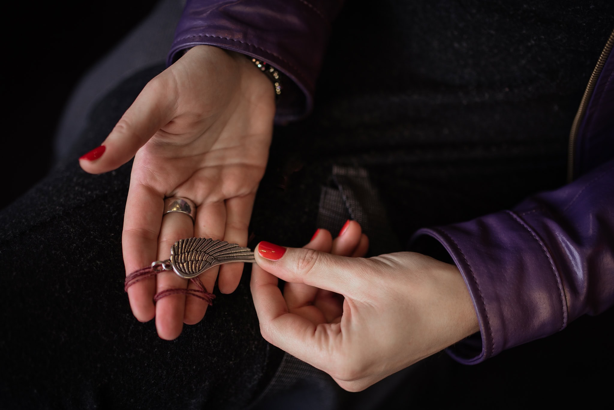 Valerie Teller's hands, adorned with red nail polish and a ring, hold a delicate silver wing charm on a crimson cord.