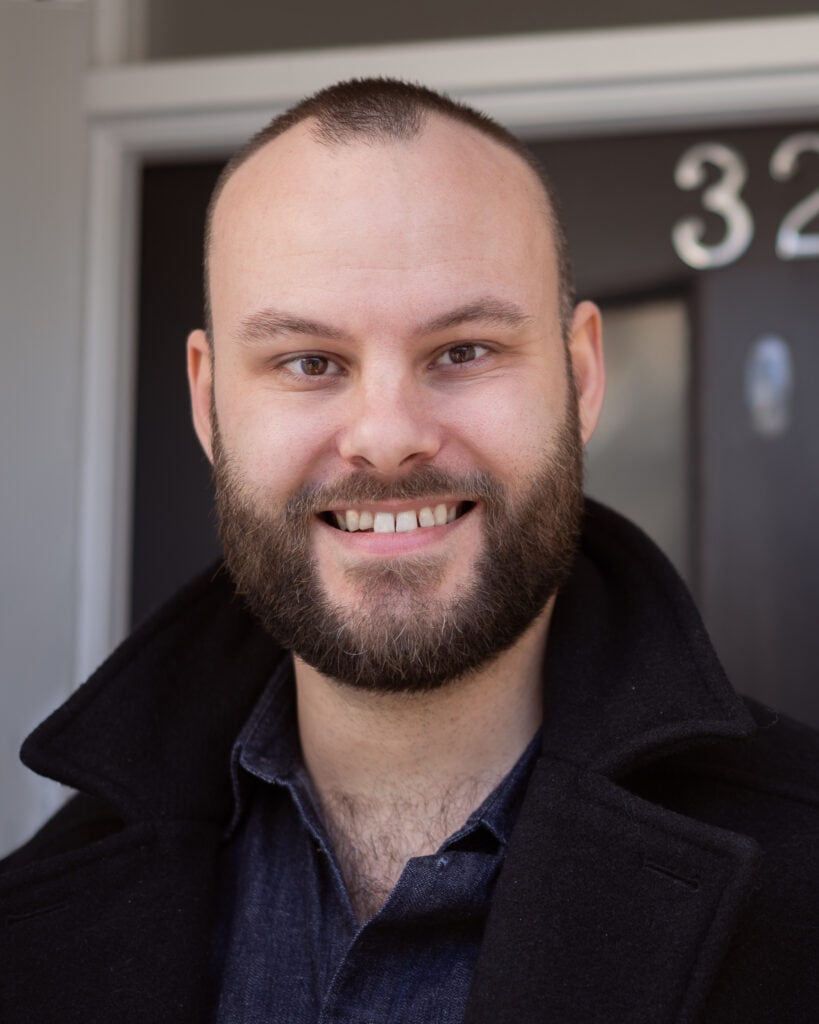 Ben Laplanche, a man with a beard, smiles in a black coat outside a building with the number 32.