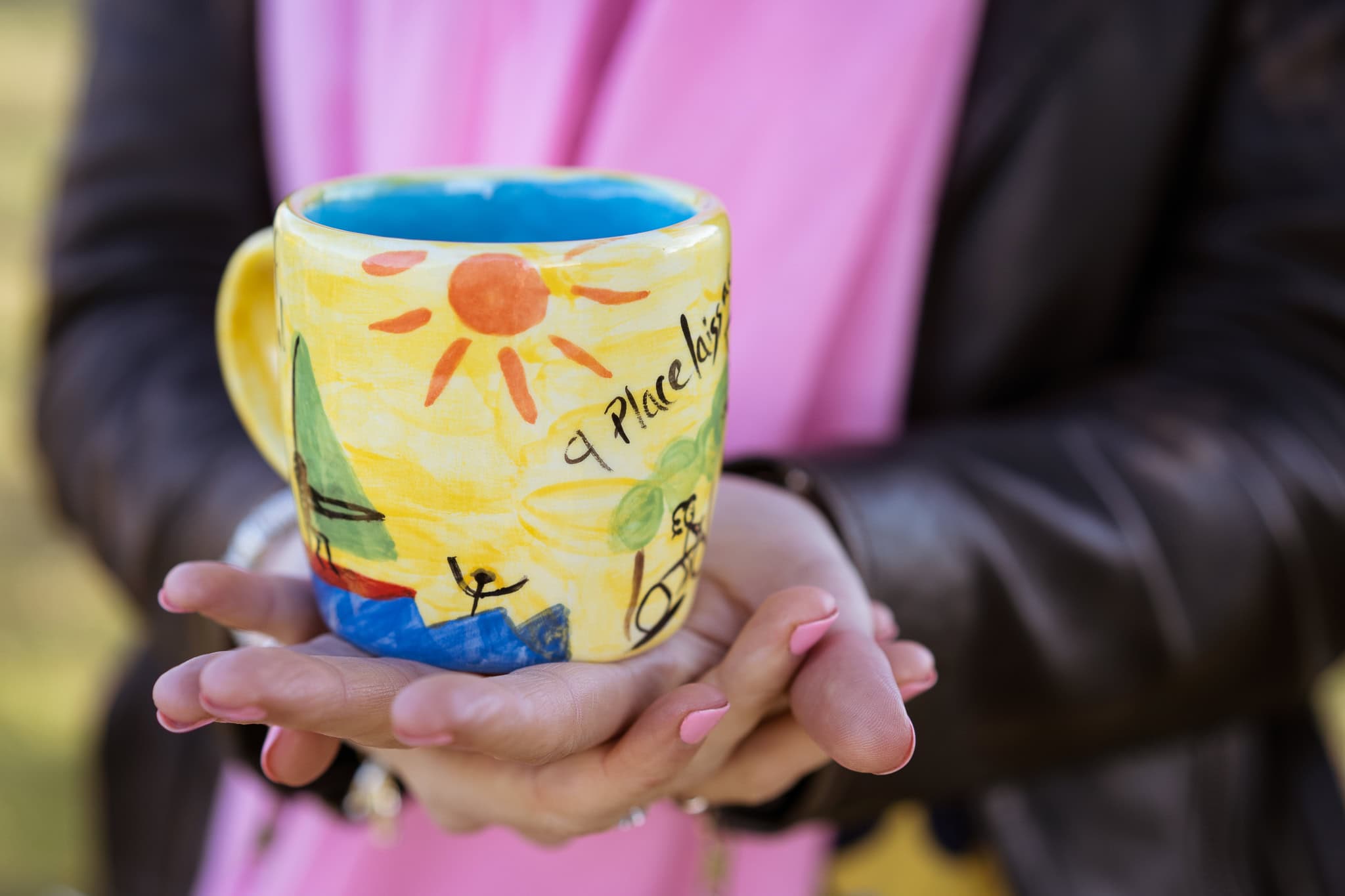 Jo Garside holds a colourful, hand-painted mug featuring a sun and a stick figure, with a pink scarf and brown jacket.