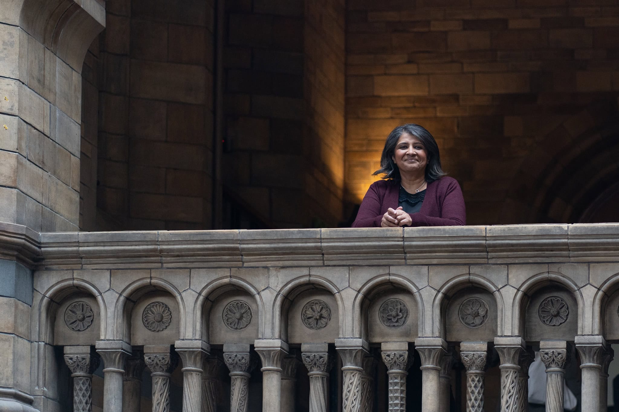 Anita Patel, a speaker, stands on a balcony in a grand building, bathed in warm light, captured by a London brand photographer.