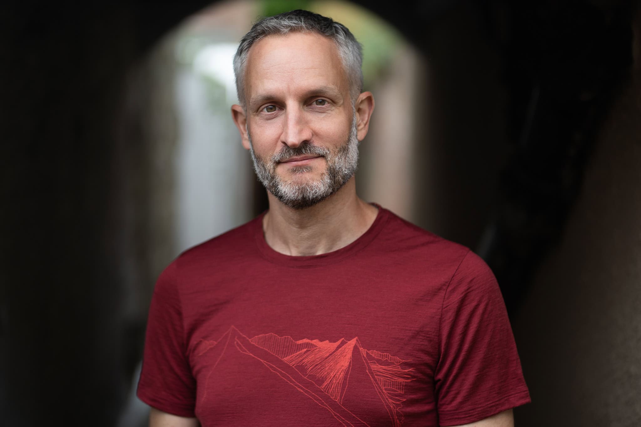 Andrew Willson, a man with grey hair and a beard, wears a red t-shirt with a mountain graphic, set against a dark, arched background.