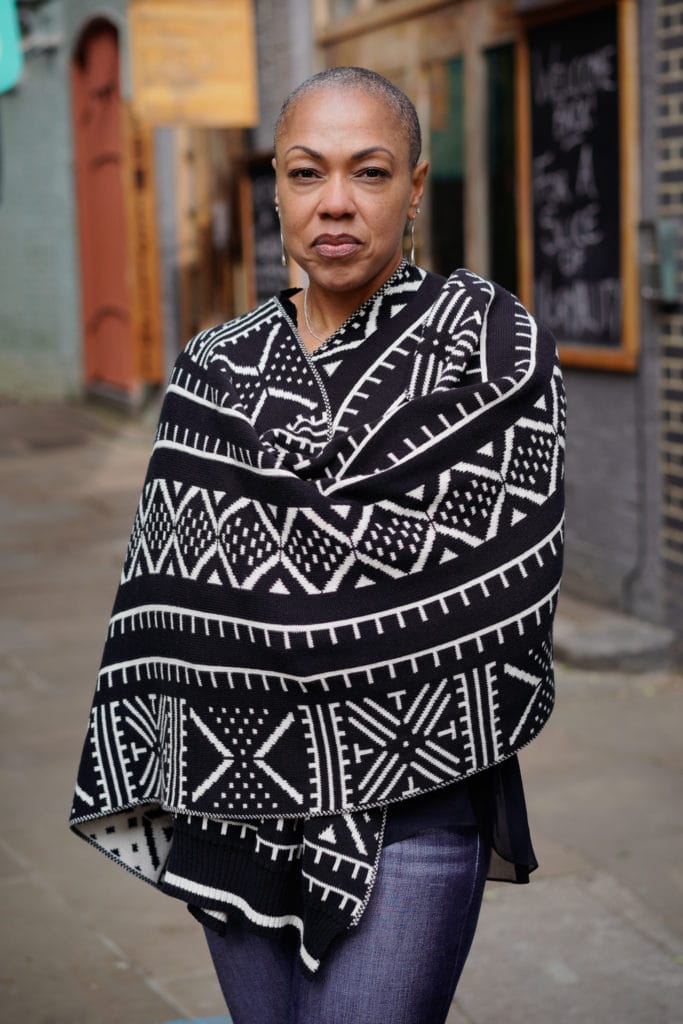Sharlene Jones, a Black woman with a shaved head, stands confidently wearing a striking monochrome patterned shawl in a London street setting.