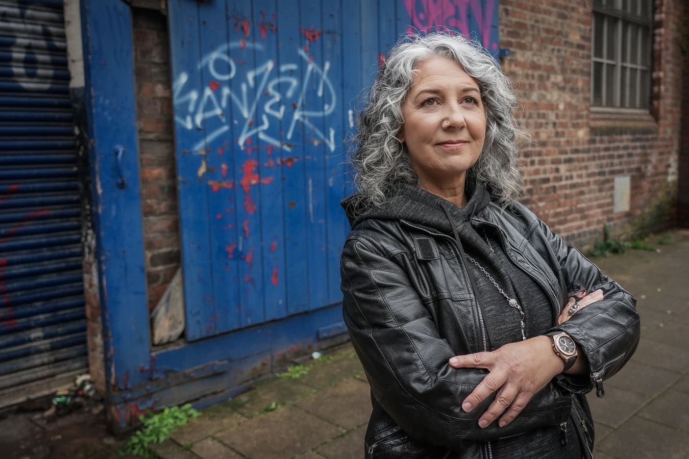 Zena Me, a confident woman with grey curly hair, in a leather jacket, stands with arms crossed in front of a graffiti-covered blue door.