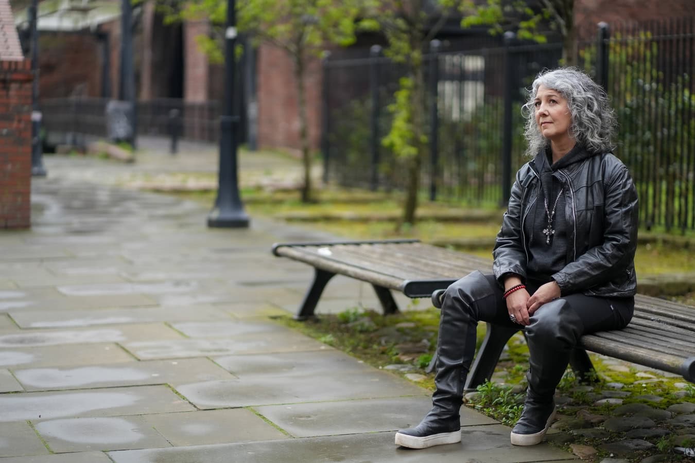 Zena Me, a woman with grey curly hair, sits on a wooden bench in a London street setting, wearing a leather jacket. Brand photography for coaches.