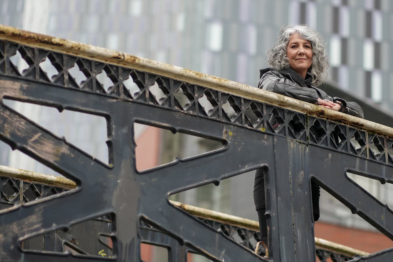Zena Me, a confident woman with grey curls, leans on a bridge railing in London, captured by a London brand photographer.