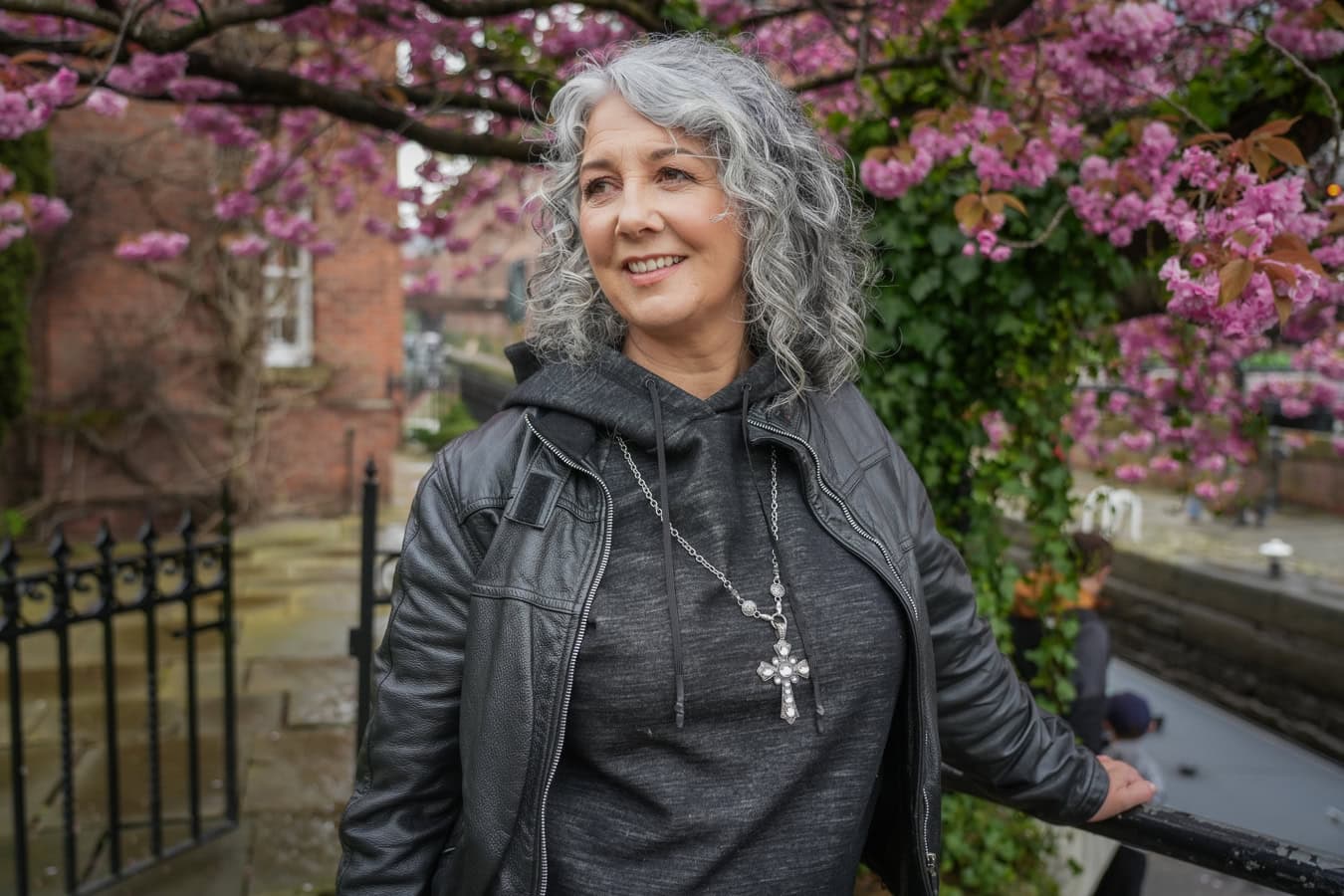 Zena Me, a confident woman with grey curly hair, smiles warmly in London, framed by pink cherry blossoms. London brand photographer.