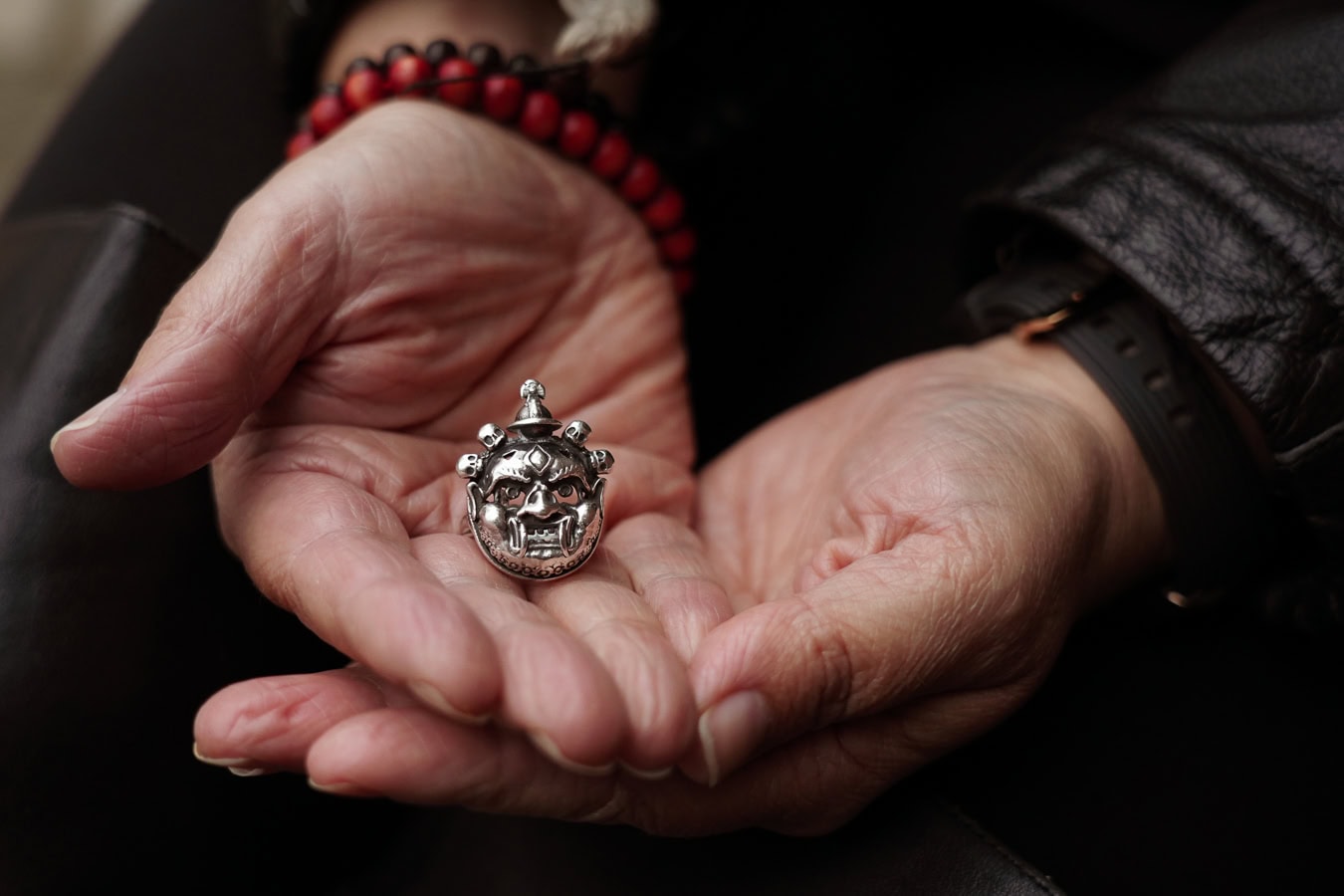 A silver, detailed mask ring rests in the open palm of a hand, with a red beaded bracelet visible.