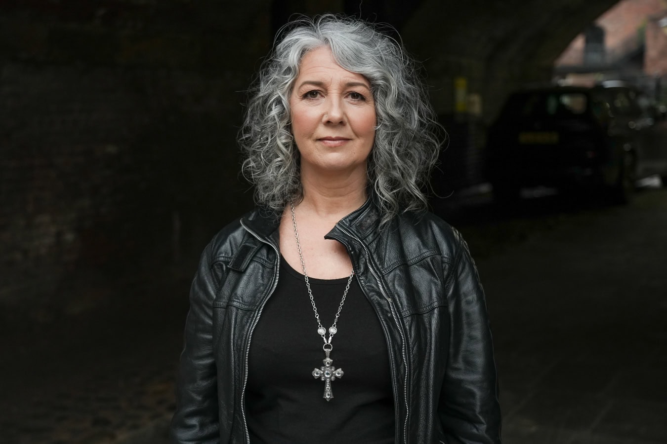 Zena Me, a confident woman with grey curly hair, in a black leather jacket and silver cross necklace, photographed by a London brand photographer.