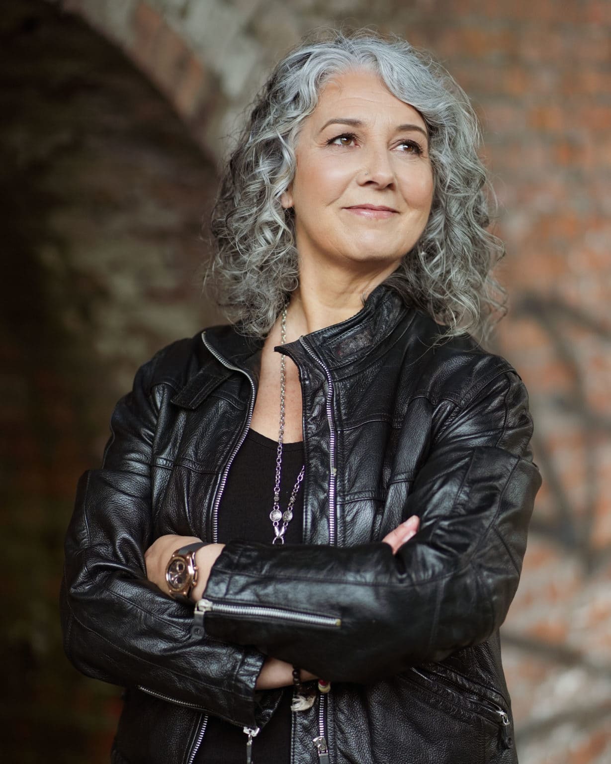Zena Me, a confident woman with grey curls, wears a black leather jacket and smiles warmly, captured by a London brand photographer.