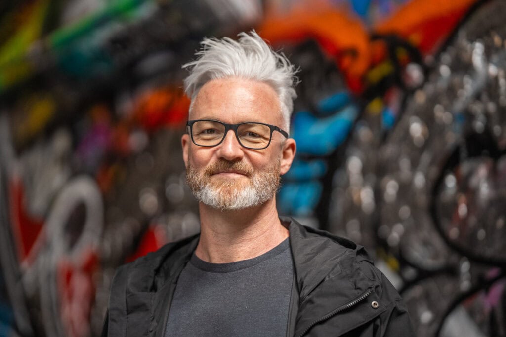 Phil Askew, London brand photographer, captured in a warm, authentic portrait with colourful graffiti background.