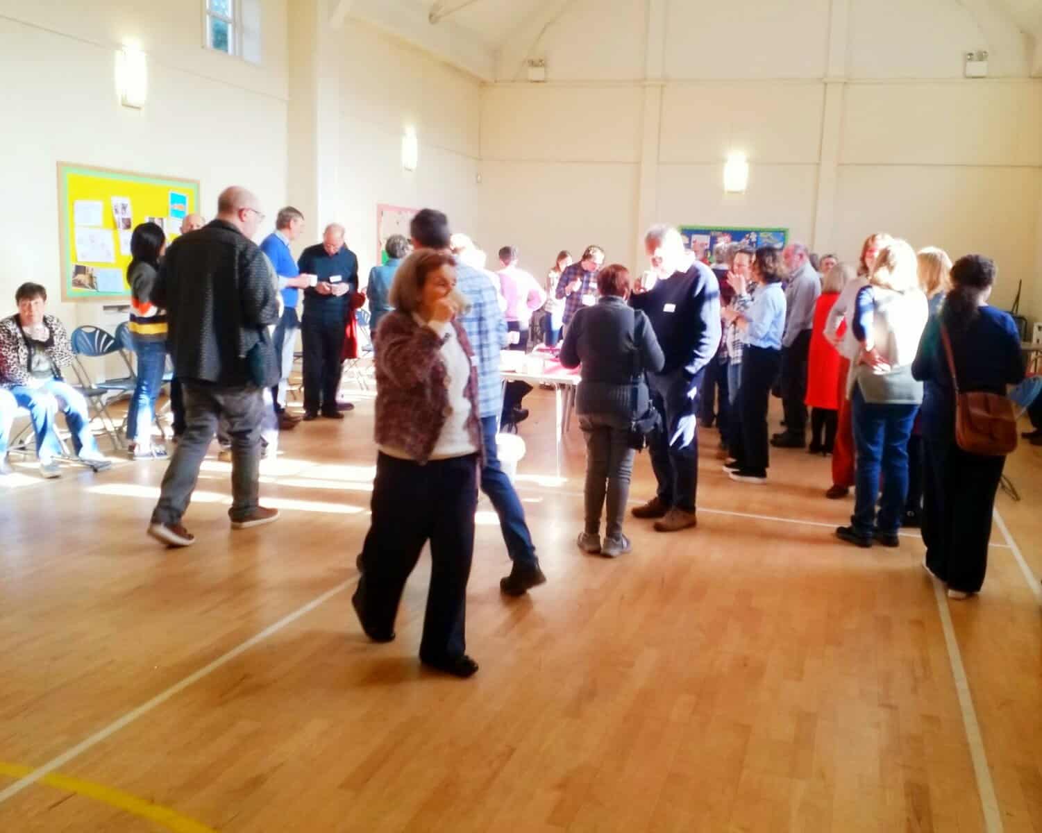People gathered at a community event held by Oriana Choir Macclesfield in a spacious hall with natural sunlight.
