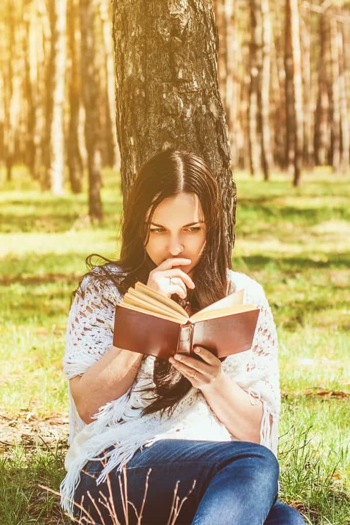woman sits against tree in the woods reading a book