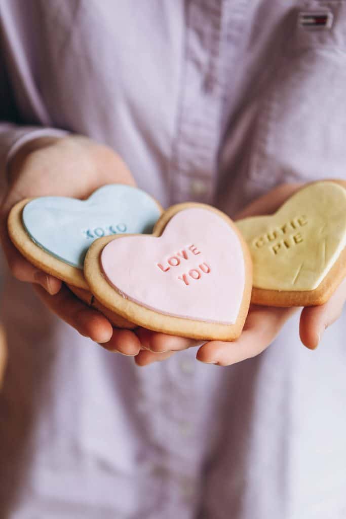 Hands holding three heart-shaped Valentine's Day sugar cookies with pastel frosting featuring messages including 'Love You' in pink, 'XOXO' in blue, and 'Cutie Pie' in yellow. Close-up photo showing delicate lettering and soft romantic lighting.