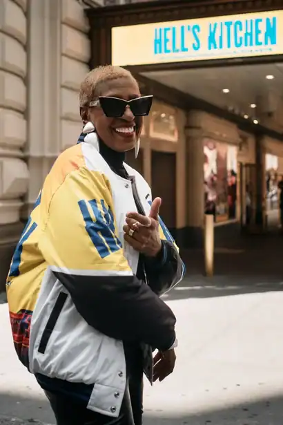Vibrant woman in stylish streetwear smiling in front of Hell's Kitchen restaurant.