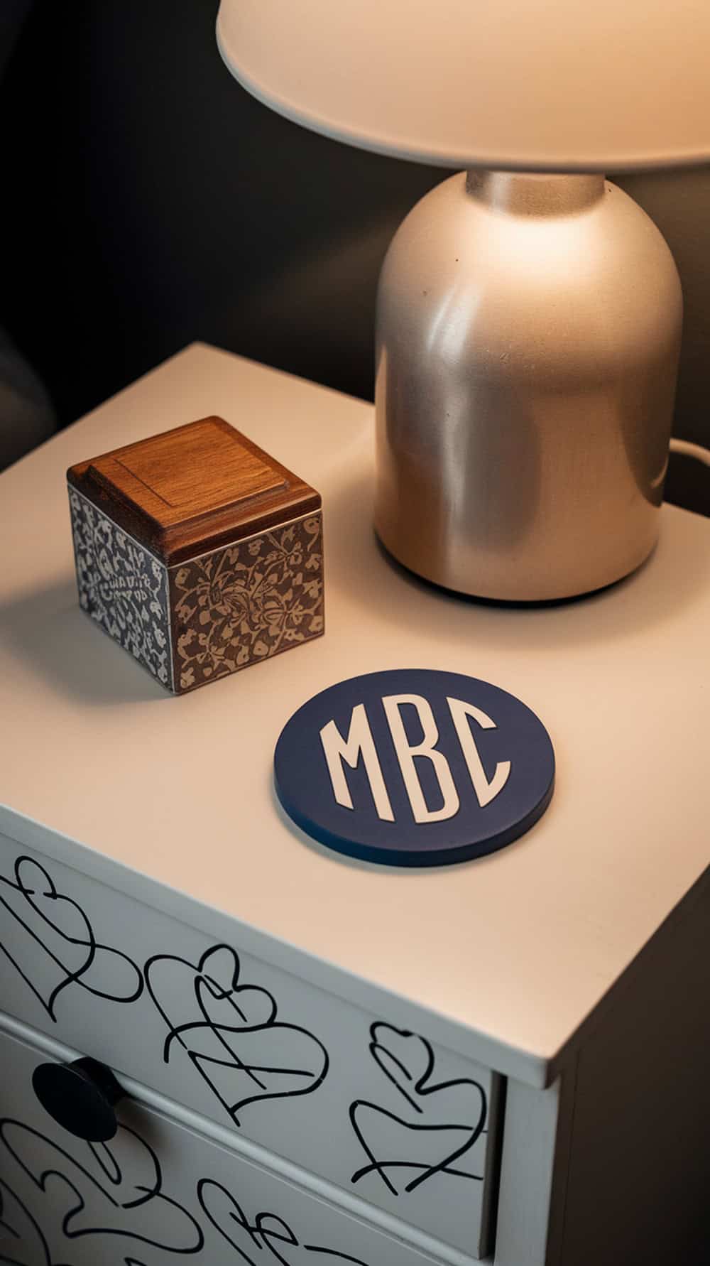 A nightstand featuring a monogrammed coaster, a decorative box, and a lamp.