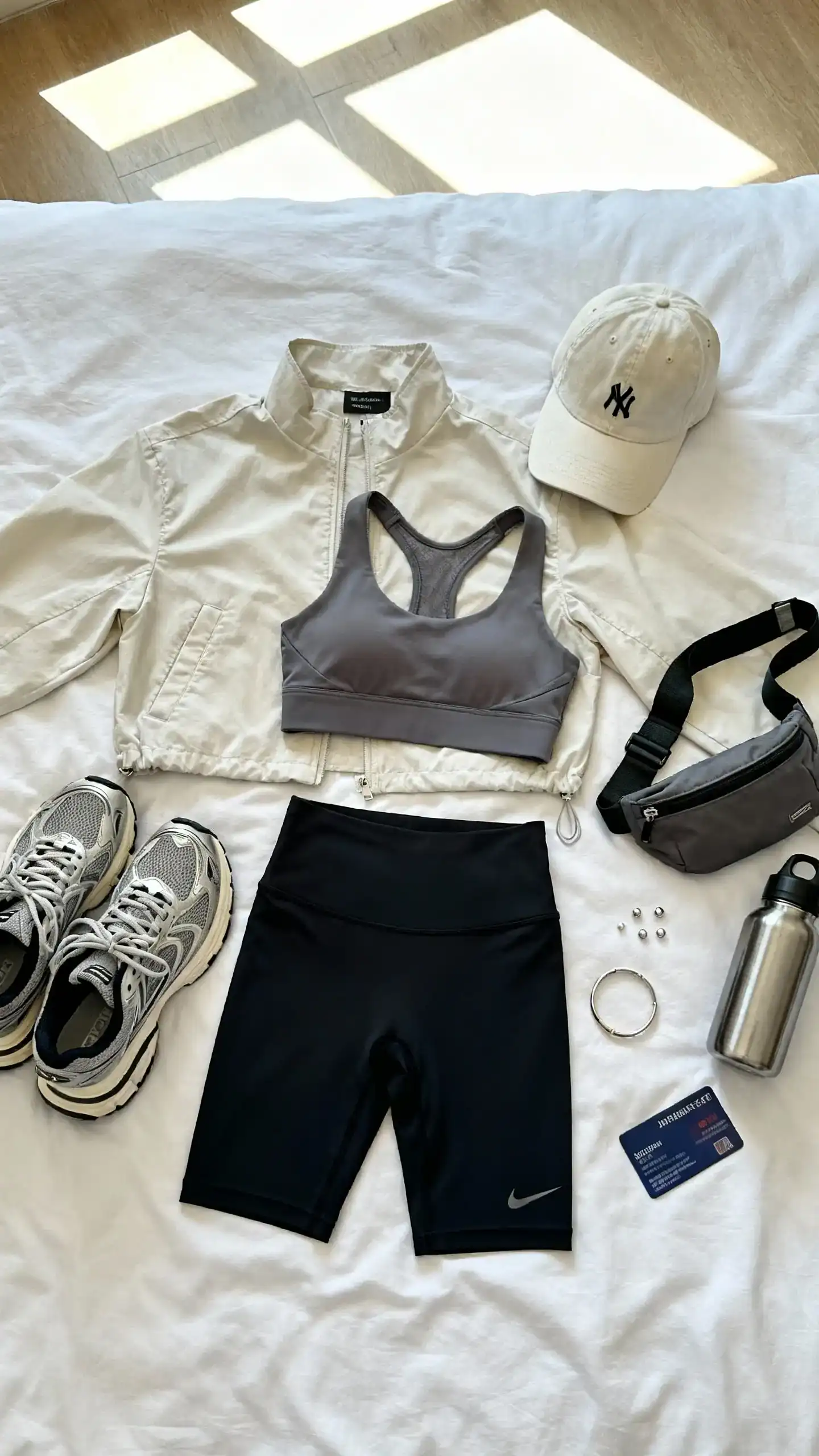 Flat lay overhead view of a light shacket, high-waisted bike shorts, supportive sports bra, chunky sneakers, baseball cap, practical belt bag, simple studs, and a slim bracelet on a white bedspread; add a reusable water bottle and gym pass, bright natural window light, no person visible, organized composition, iPhone photo aesthetic.