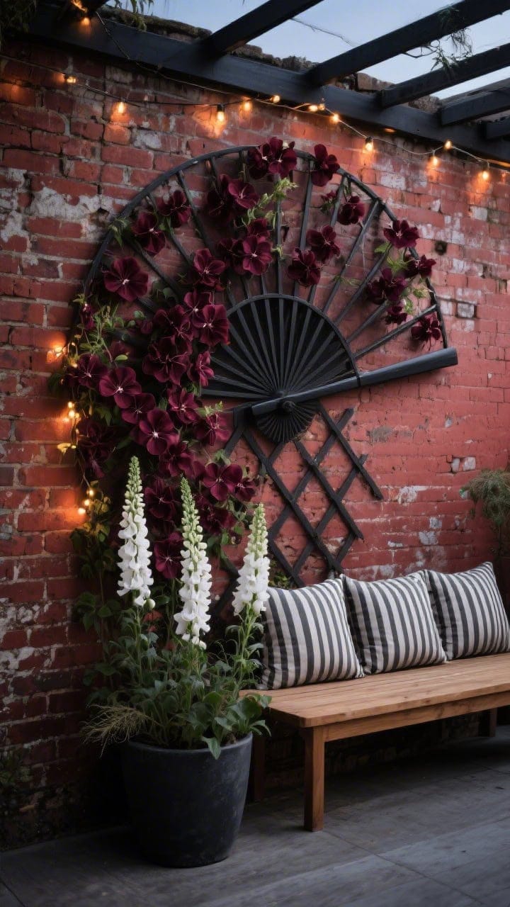 Photorealistic medium shot, straight-on view of a blackened iron fan trellis leaning against an aged red brick wall; deep burgundy clematis sweeping across the fan’s spokes; at the base, white foxgloves rising beside charcoal planters; a teak bench with stripey cushions softens the scene; warm fairy lights draped low along the trellis arms add a moody glow; textures emphasized: rough brick, smooth metal, feathery flowers; color vibe red brick, black iron, inky blooms, crisp white; dusk lighting, no people