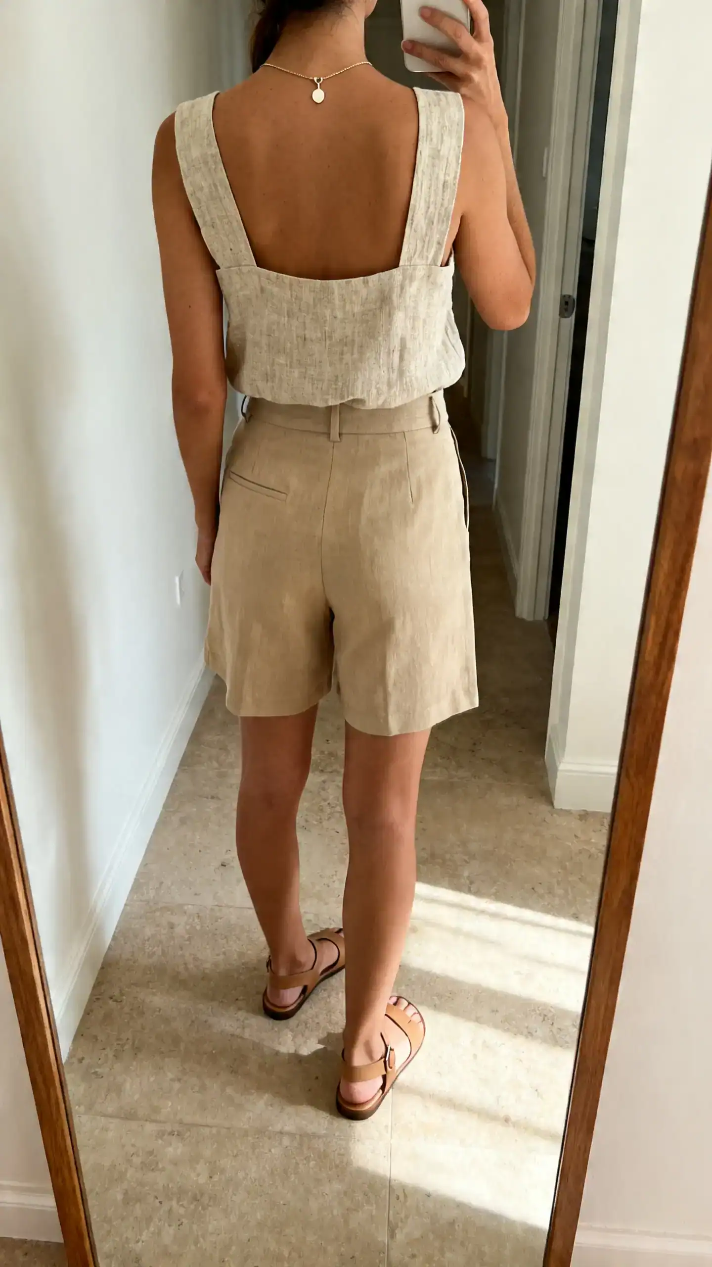 iPhone back-view selfie of a woman wearing a linen tank with elevated wide straps, tailored beige shorts, minimalist leather sandals, delicate pendant necklace, face not visible, hallway full-length mirror, natural afternoon light, iPhone photo quality