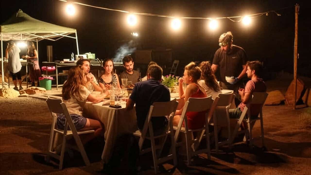 קבוצה יושבת סביב שולחן אוכל בערב במדבר תחת שרשרת נורות לצד מאהל