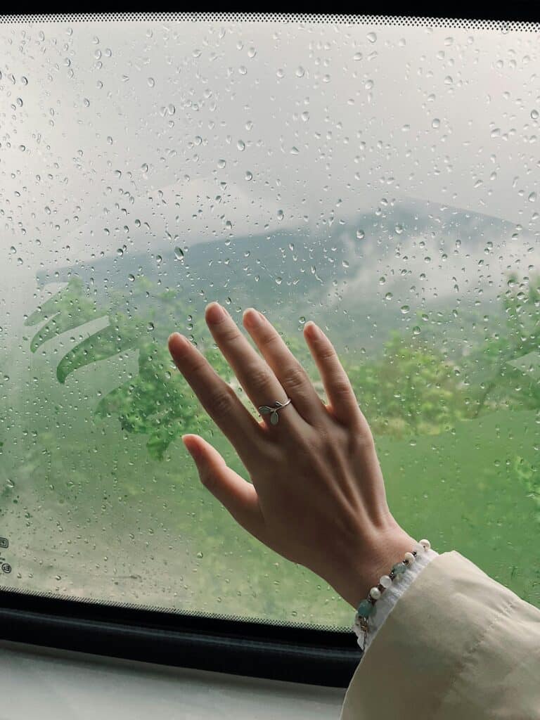 A hand wearing a ring touches a raindrop-covered window overlooking lush greenery.