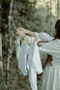 Pregnant woman hanging baby clothes on a clothesline in a lush green outdoor setting.
